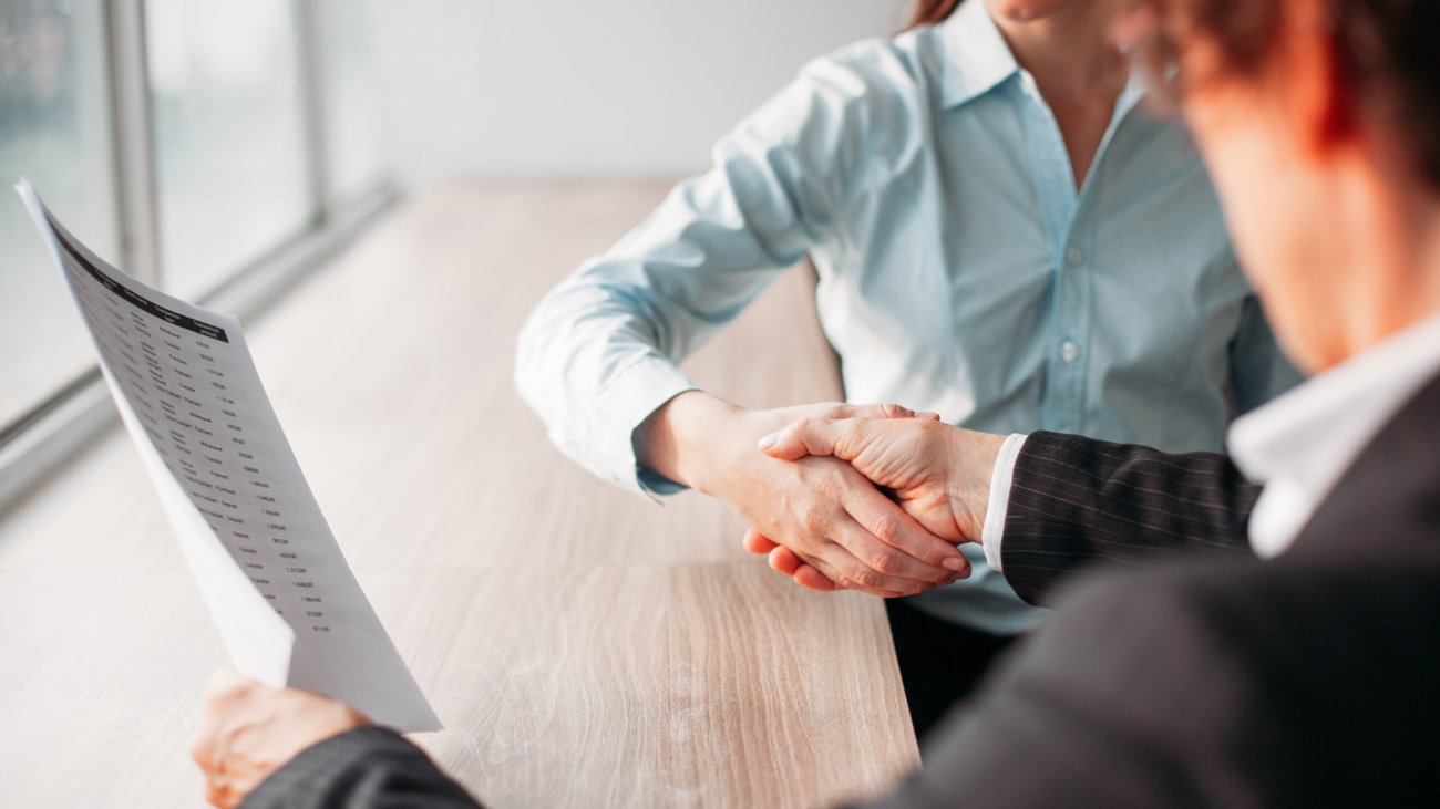 Unrecognizable businesswoman holding document and shaking hand of business partner after signing of contract. They sitting at table. Negotiation concept